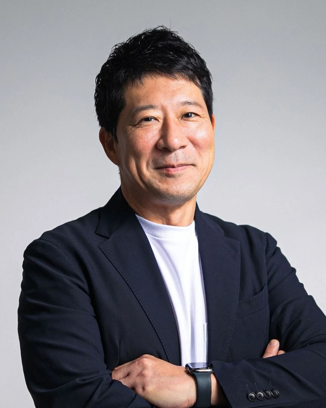 白いTシャツとダークカラーのジャケットを着用し、腕を組んで微笑むアジア人の中年男性のポートレート。ビジネスシーンにも合う落ち着いた雰囲気で、信頼感のある表情が印象的です。