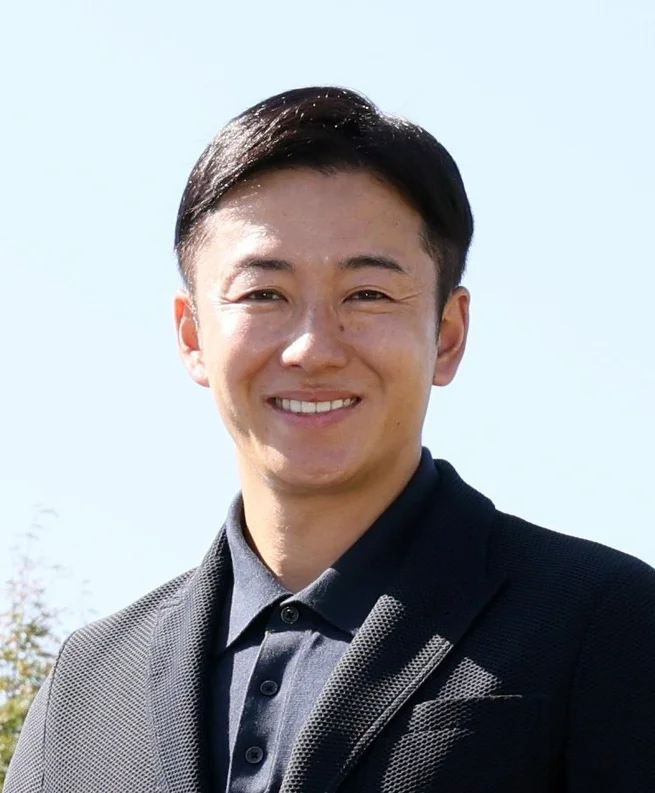 青空の下、ダークカラーのポロシャツとジャケットを着用した斎藤佑樹氏が、穏やかな笑顔でカメラを見ているポートレート。