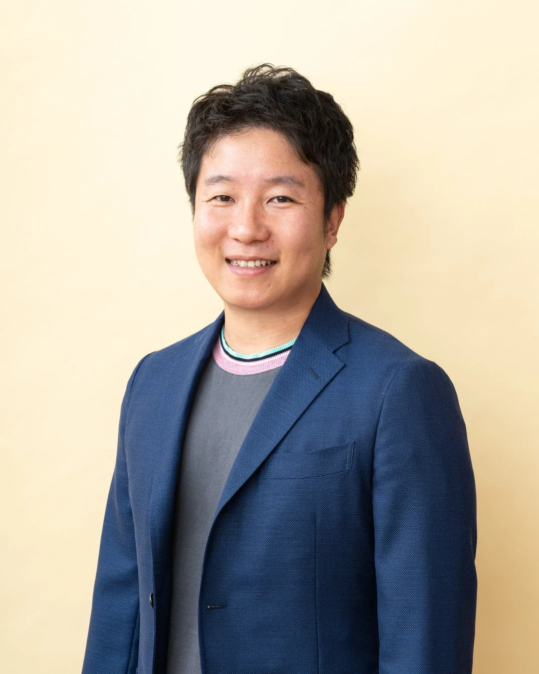 明るい黄色の背景の前で笑顔を見せるアジア人男性のポートレートです。男性は青いジャケットとグレーのTシャツを着用しており、首元にはカラフルなボーダー柄が見えます。