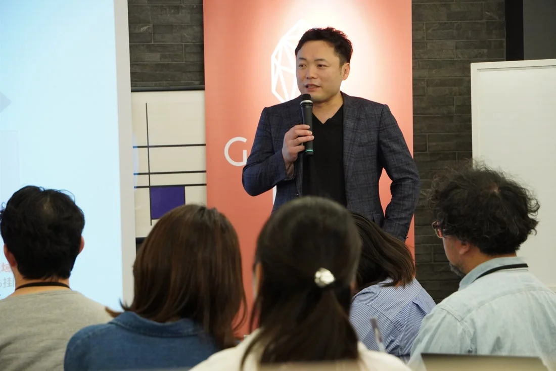 マイクを持った男性が聴衆に向けて講演を行っている様子