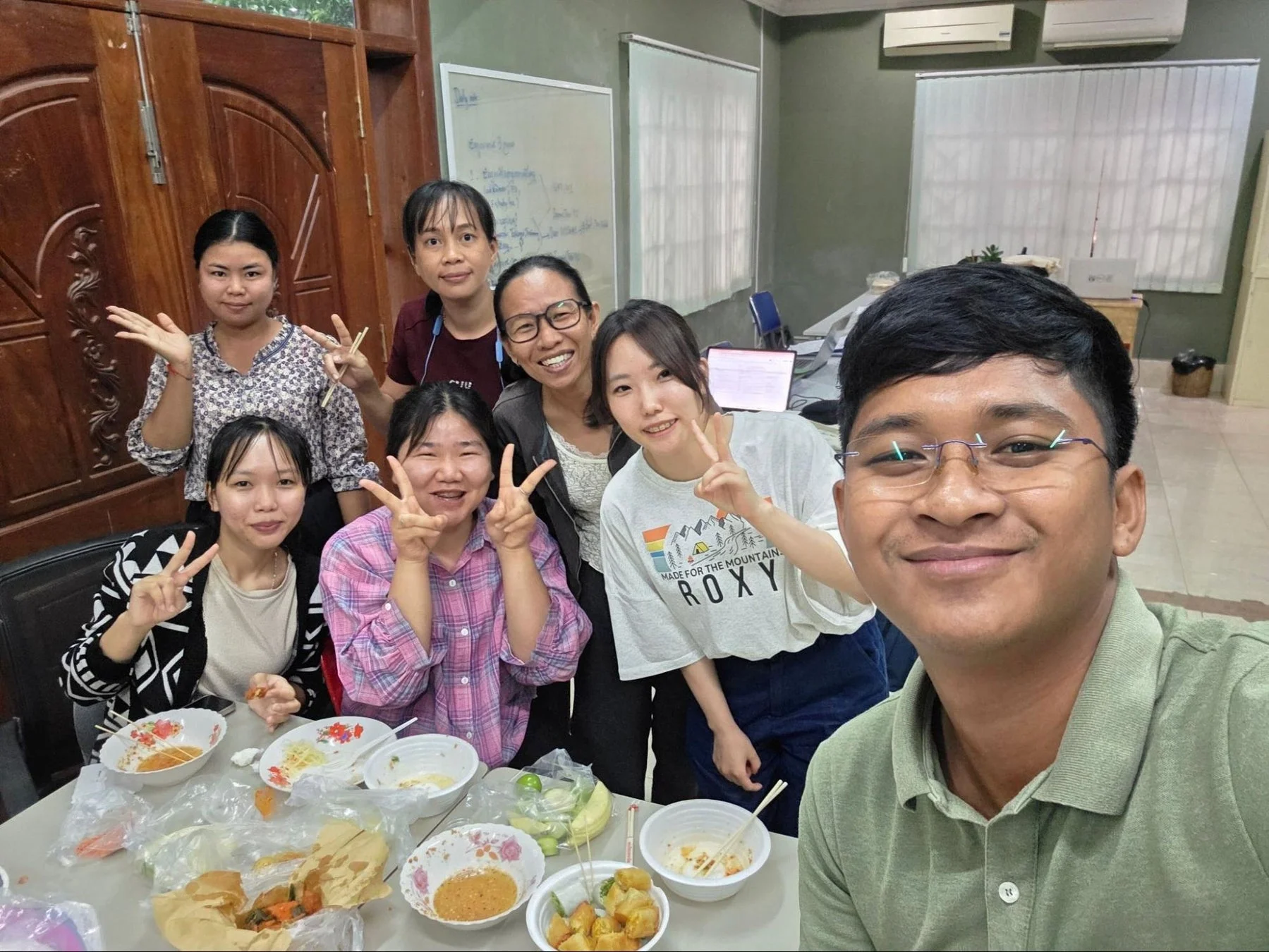 笑顔のアジア人グループが食卓を囲んでカジュアルな食事を楽しんでいる様子。友人や同僚が集まり、和やかな雰囲気の中でセルフィーを撮っている。