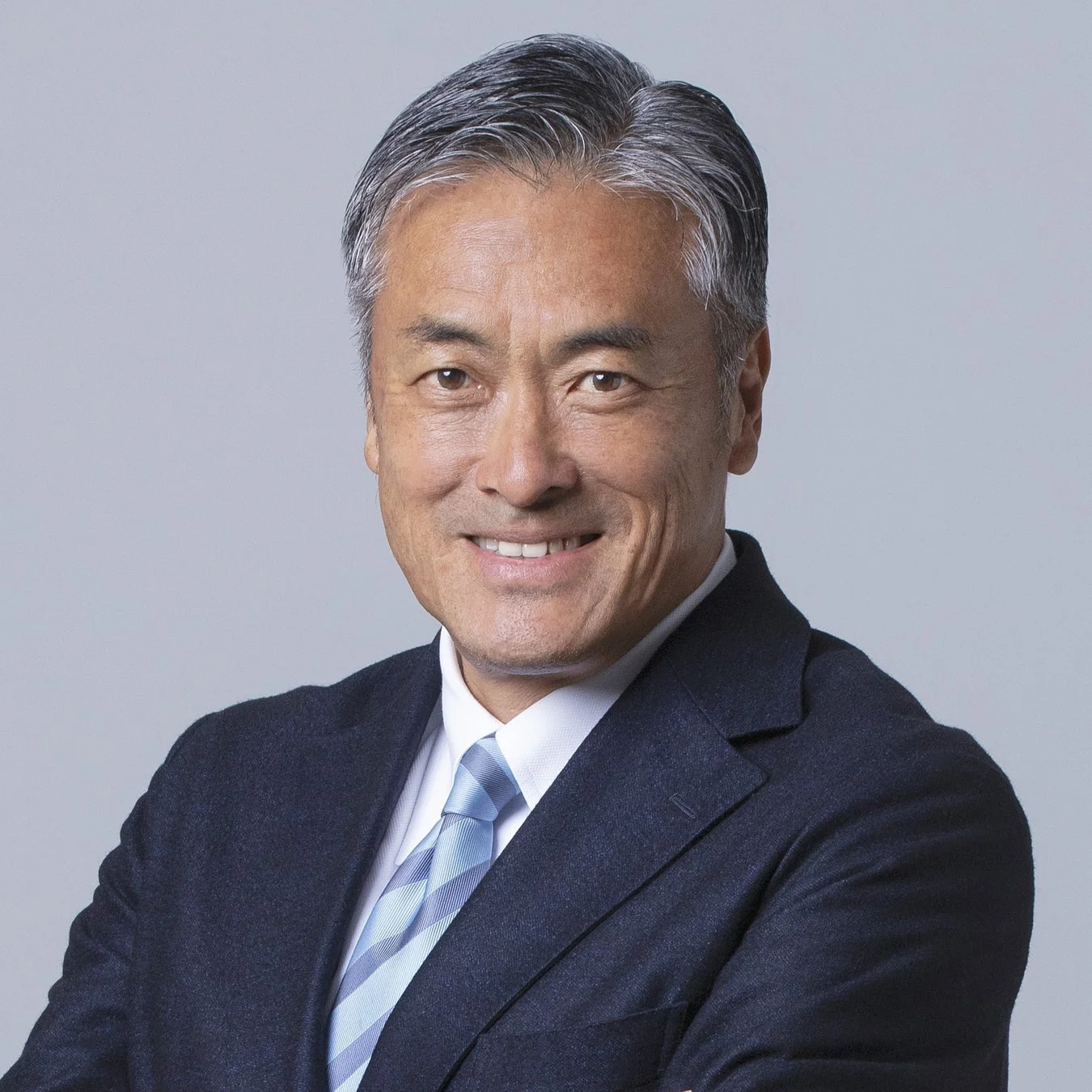 明るい灰色の背景の前で、ダークスーツと水色のストライプネクタイを着用した中高年アジア人男性のポートレートです。男性はカメラに向かって穏やかに微笑んでおり、プロフェッショナルで親しみやすい印象を与えます。