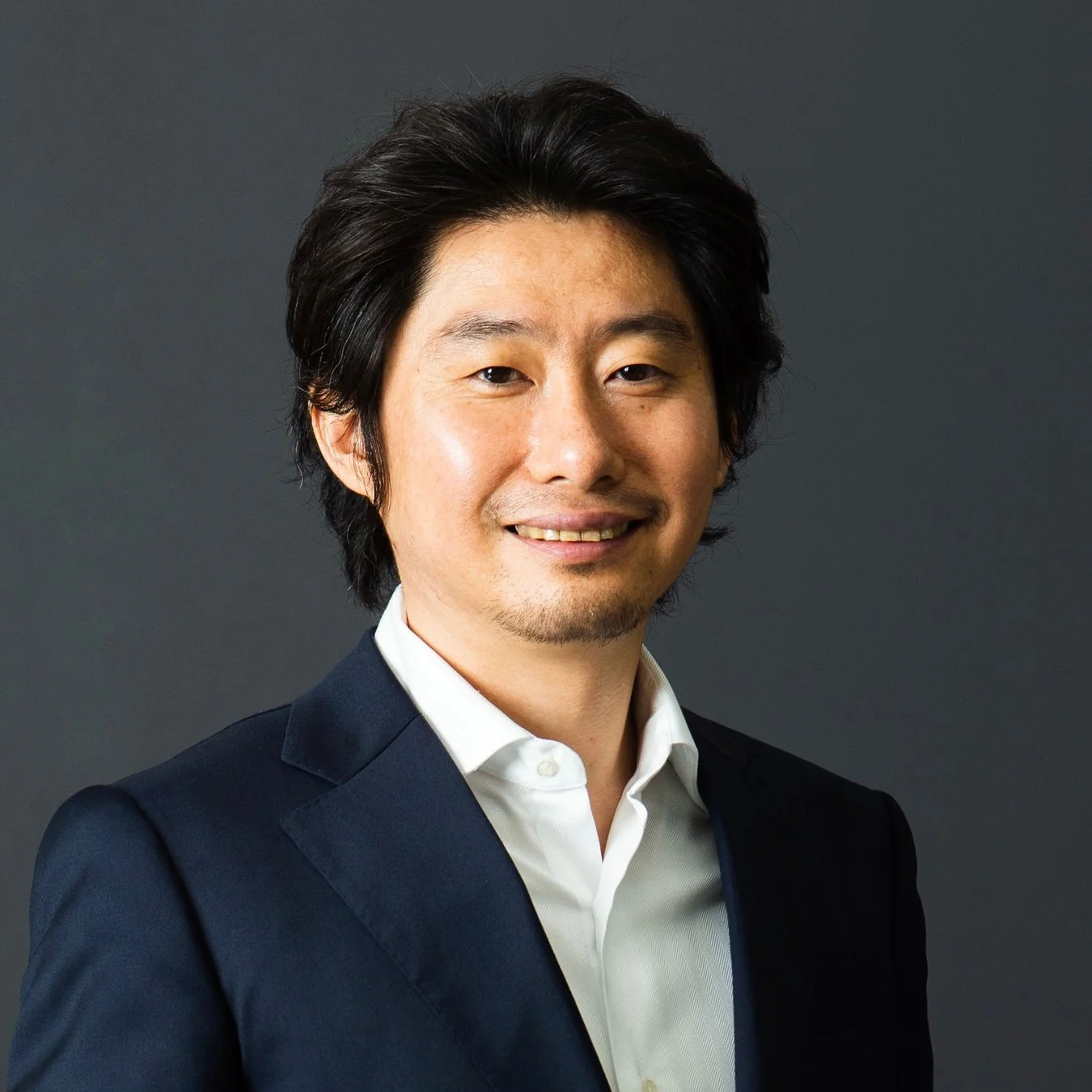暗い背景の前で、紺色のスーツジャケットと白いシャツを着用し、笑顔を見せるアジア人男性のポートレートです。親しみやすい印象を与えています。