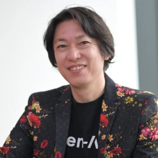 笑顔でカメラを見つめる日本の男性のポートレート。黒いTシャツの上にカラフルな花柄のジャケットを着用しており、背景は明るい色でシンプルです。