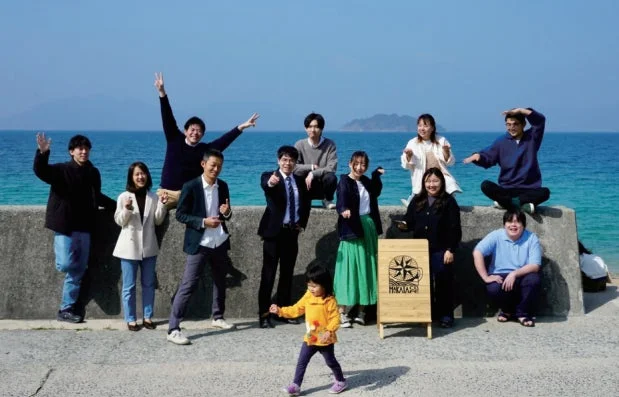 晴れた日に青い海を背景に、多くの大人と一人の子供が笑顔でポーズをとっている集合写真です。コンクリートの防波堤に座ったり立ったりしており、木製の看板も写っています。