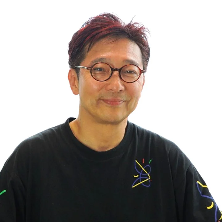 白い背景を背に、丸いメガネをかけたアジア人男性が優しく微笑んでいます。赤いメッシュの入った短髪で、カラフルな抽象画がプリントされた黒いTシャツを着ています。