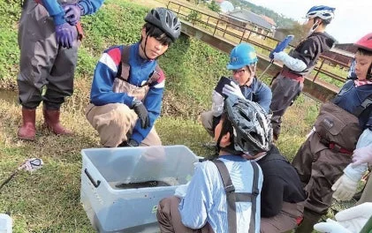 川辺で複数の人々がウェットスーツやヘルメットを着用し、水槽を囲んで何らかの活動を行っている様子です。環境学習や生態調査のようなフィールドワークをしていると推測されます。