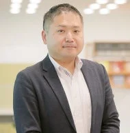 ビジネススーツを着用したアジア人男性のポートレートです。男性はカメラに向かっており、真面目な表情をしています。背景はぼかされており、屋内であることが示唆されます。