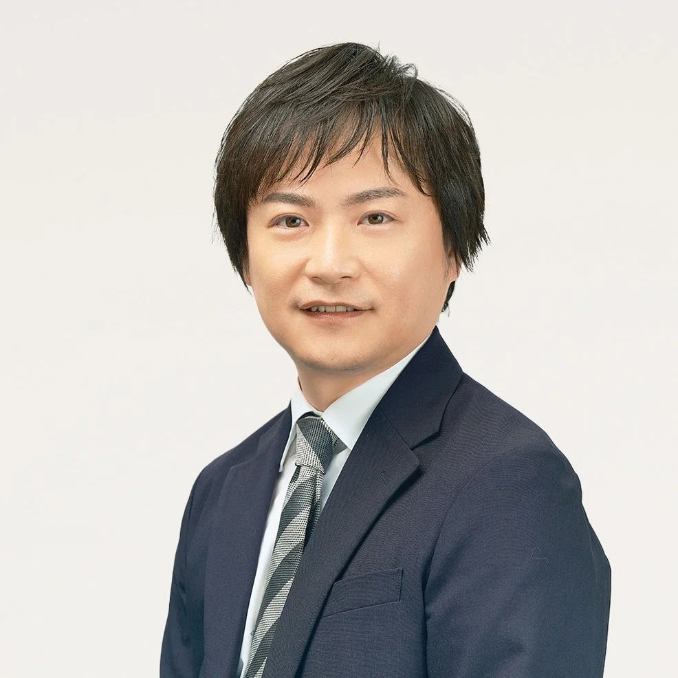 白い背景の前で、ダークスーツとストライプのネクタイを着用したアジア人男性がカメラに向かって穏やかに微笑んでいるポートレート写真です。清潔感のあるビジネスシーンに適した一枚。