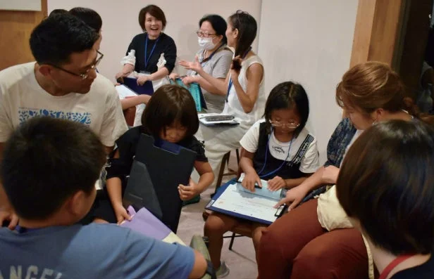 複数の大人と子供たちが屋内で集まり、何らかの活動に参加している様子。子供たちは書類やタブレットに集中し、大人たちは互いに会話したり子供たちを見守ったりしている。和やかな雰囲気の中で学習や交流が行われているようだ。