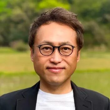 眼鏡をかけたアジア人男性のポートレート。黒いジャケットと白いシャツを着用し、穏やかな笑顔を浮かべている。背景は緑豊かな屋外がぼやけている。