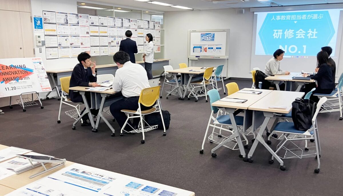 オフィスのような空間で、複数のグループがテーブルを囲んで話し合っています。スクリーンには「人事教育担当者が選ぶ 研修会社 NO.1」と表示され、左側には「LEARNING INNOVATION AWARDS 2025」のポスターが見えます。研修やビジネスイベントの様子を捉えた画像です。