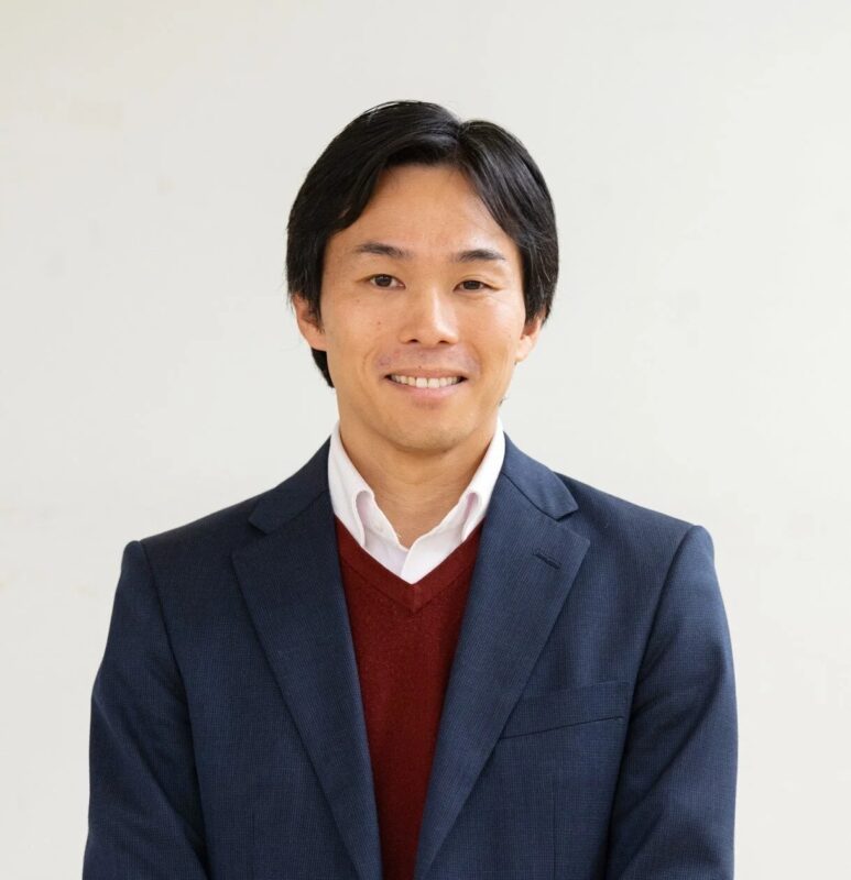 白い背景の前で、紺色のスーツと赤いVネックセーターを着用し、笑顔を見せるアジア人男性のポートレートです。清潔感があり、ビジネスシーンやプロフィール写真に適した一枚です。