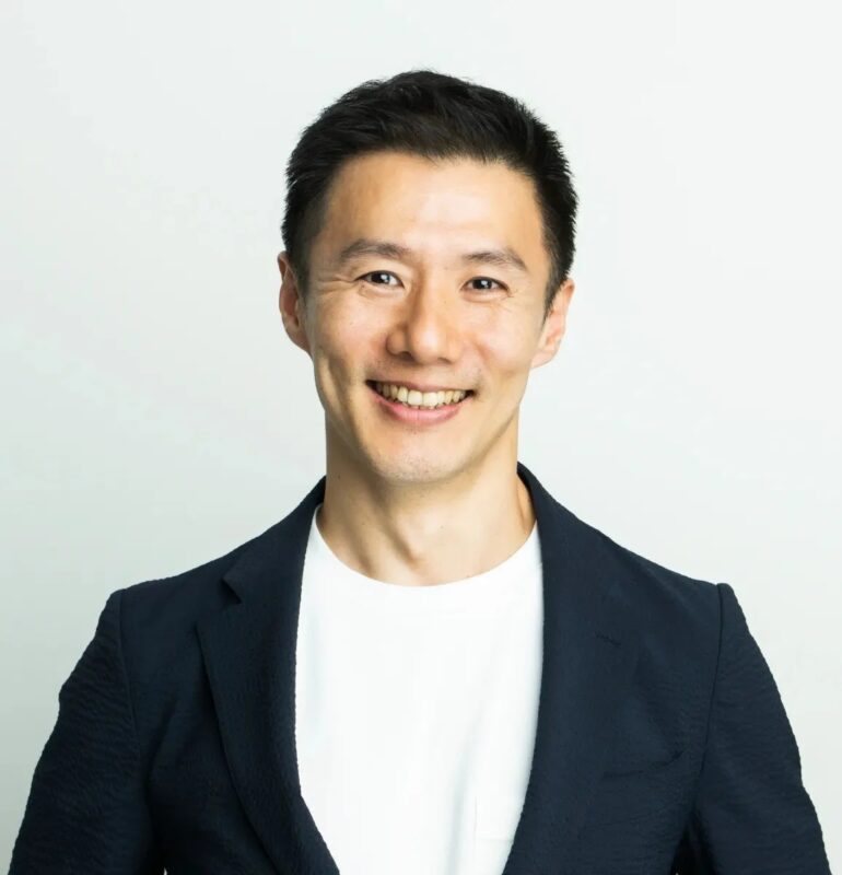 白い背景の前で、黒いジャケットと白いTシャツを着たアジア人男性がカメラに向かって笑顔を見せているポートレート写真