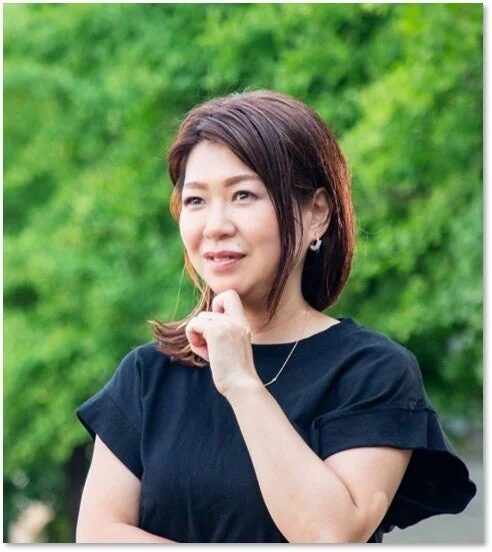 緑豊かな背景をバックに、黒いTシャツを着た女性が顎に手を当てて優しく微笑んでいるポートレートです。自然光が当たり、穏やかな雰囲気が漂っています。