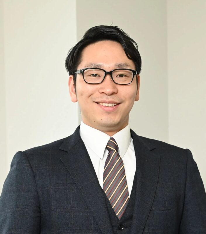 眼鏡をかけたアジア人男性が、ダークスーツとストライプのネクタイを着用し、白い背景の前で笑顔を見せている上半身のポートレートです。プロフェッショナルで親しみやすい印象を与えます。