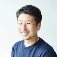 白い背景の前で笑顔を見せる、短髪でヒゲのある日本人男性のポートレートです。紺色のTシャツを着用しており、親しみやすい印象を与えます。