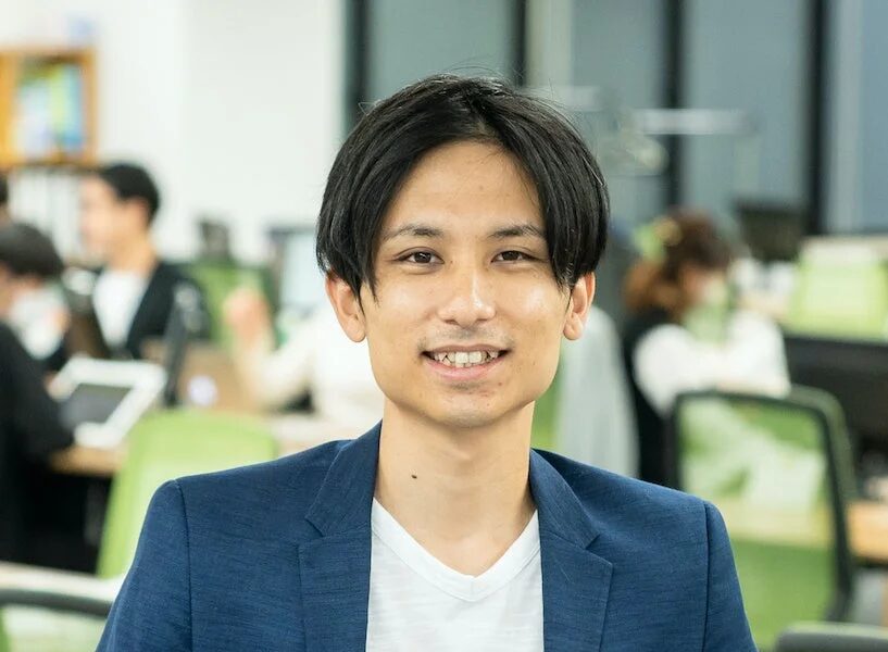 オフィスで青いジャケットと白いTシャツを着た若い男性が、カメラに向かって笑顔で写っているポートレート写真