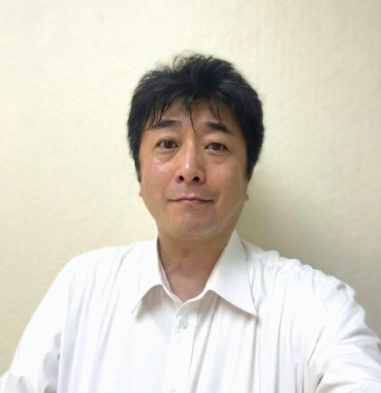 白いシャツを着たアジア人男性のポートレートです。カメラをまっすぐ見ており、背景はシンプルな壁です。