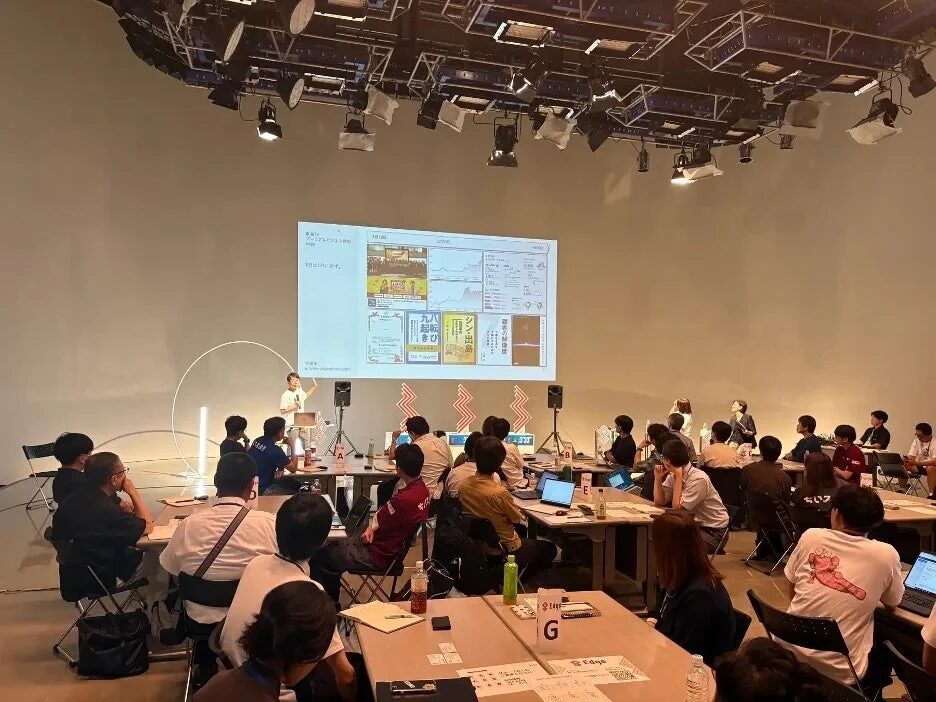 広々とした会場でプレゼンテーションが行われています。登壇者がスクリーンを指して説明し、多くの参加者がテーブルに着席して熱心に耳を傾けています。スクリーンには「デジタルビジネスの推進」といった文字が表示されています。