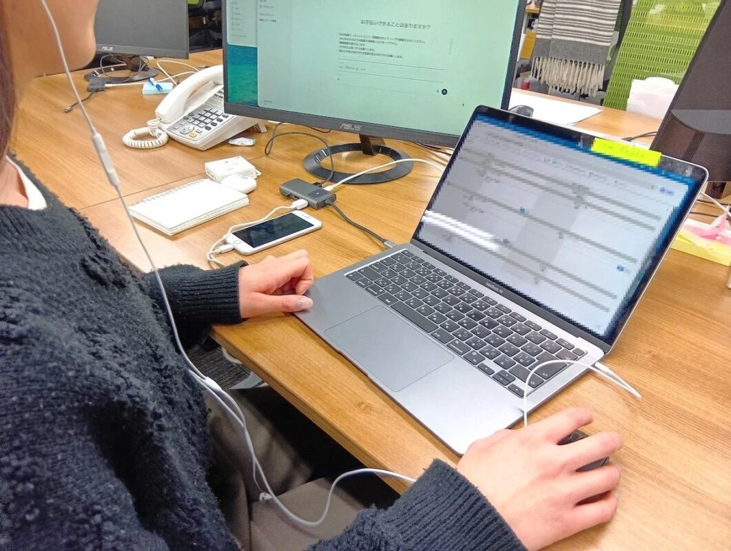 オフィスで働く人物がデスクに向かい、ヘッドホンを着用してMacBook Airと外部モニターを使用している様子