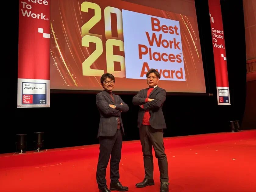 2026 Best Work Places Awardのロゴが映し出されたスクリーンを背景に、2人の男性が赤いステージに立っている。