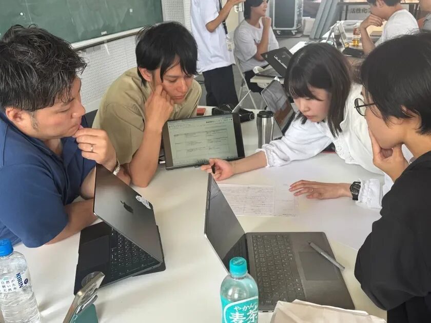 数人の男女がテーブルを囲み、ノートパソコンやタブレットを使って共同作業や議論をしている様子です。