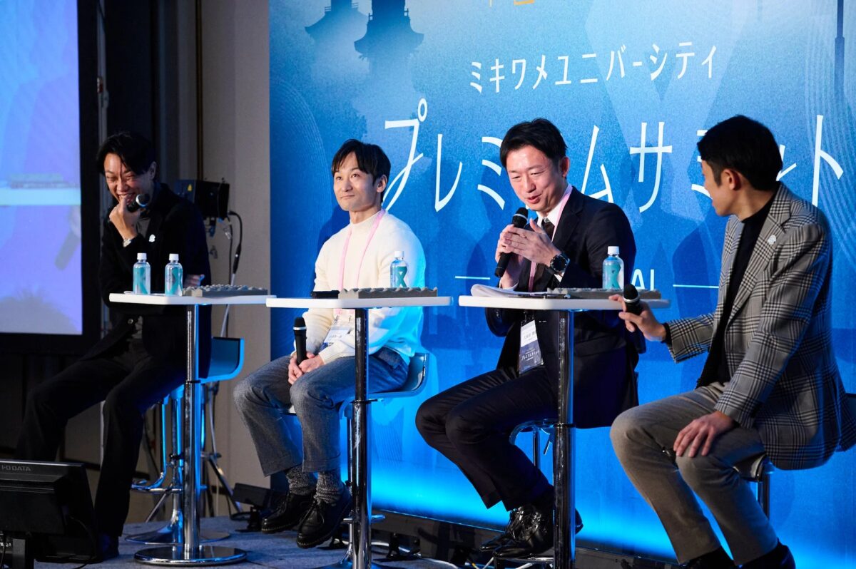 青い背景のステージで複数の男性がマイクを手に、パネルディスカッション形式で話し合っている様子。