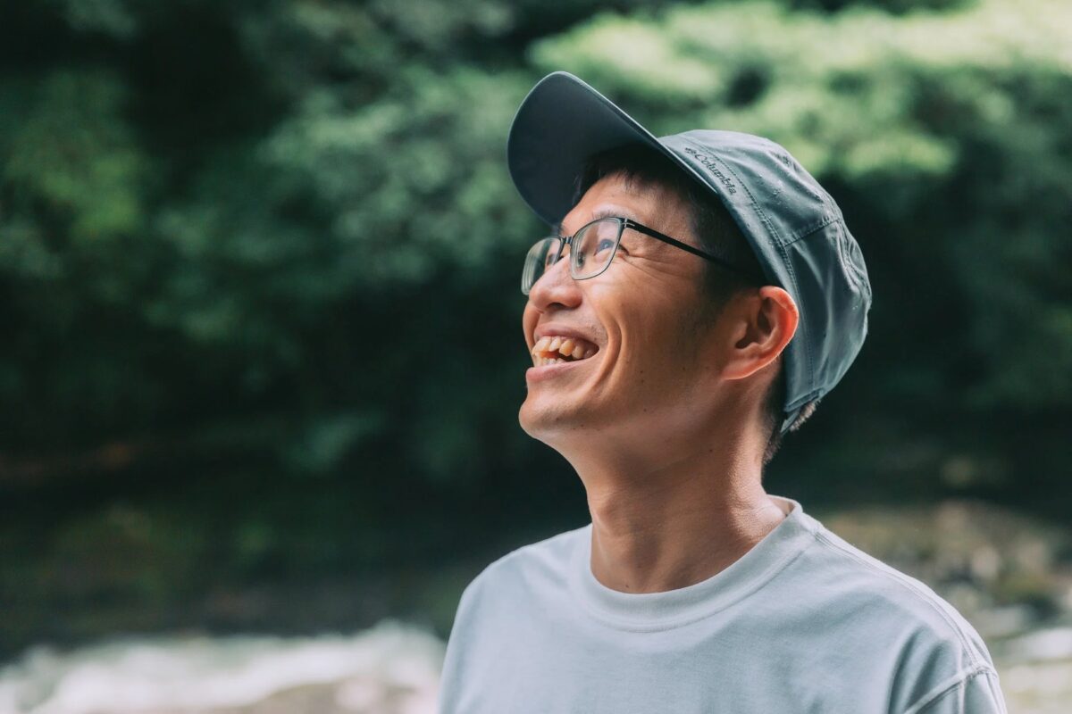 緑豊かな自然の中で、メガネと帽子をかぶった男性が満面の笑みで上を見上げています。