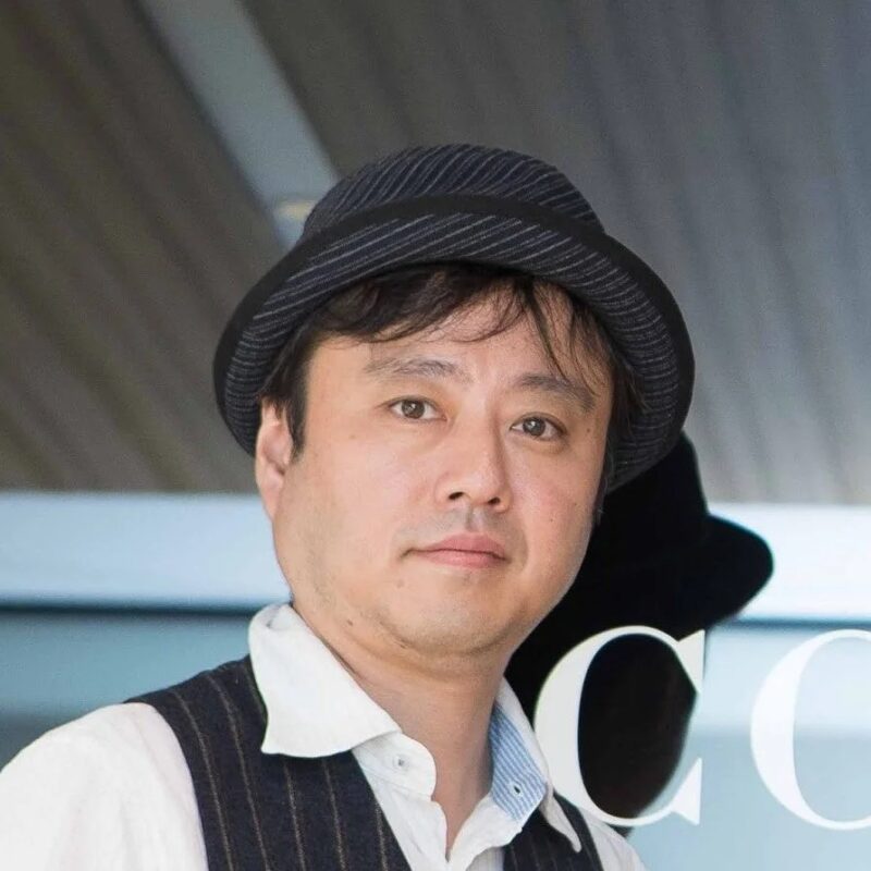 ストライプのフェドラ帽とピンストライプのベストを着たアジア人男性のポートレートです。男性はカメラをまっすぐ見ており、落ち着いた表情をしています。