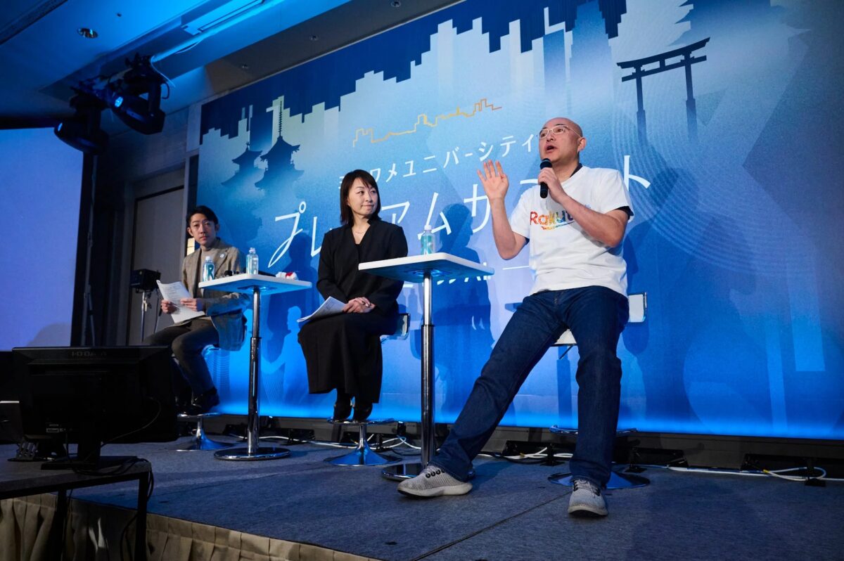 イベント会場のステージで3人の登壇者がパネルディスカッションを行っている。背景には日本の都市景観や伝統的な建築物がデザインされ、「コミュニティユニバーシティ プレミアムカンファレンス」の文字が確認できる。楽天のTシャツを着た男性がマイクで発言中。