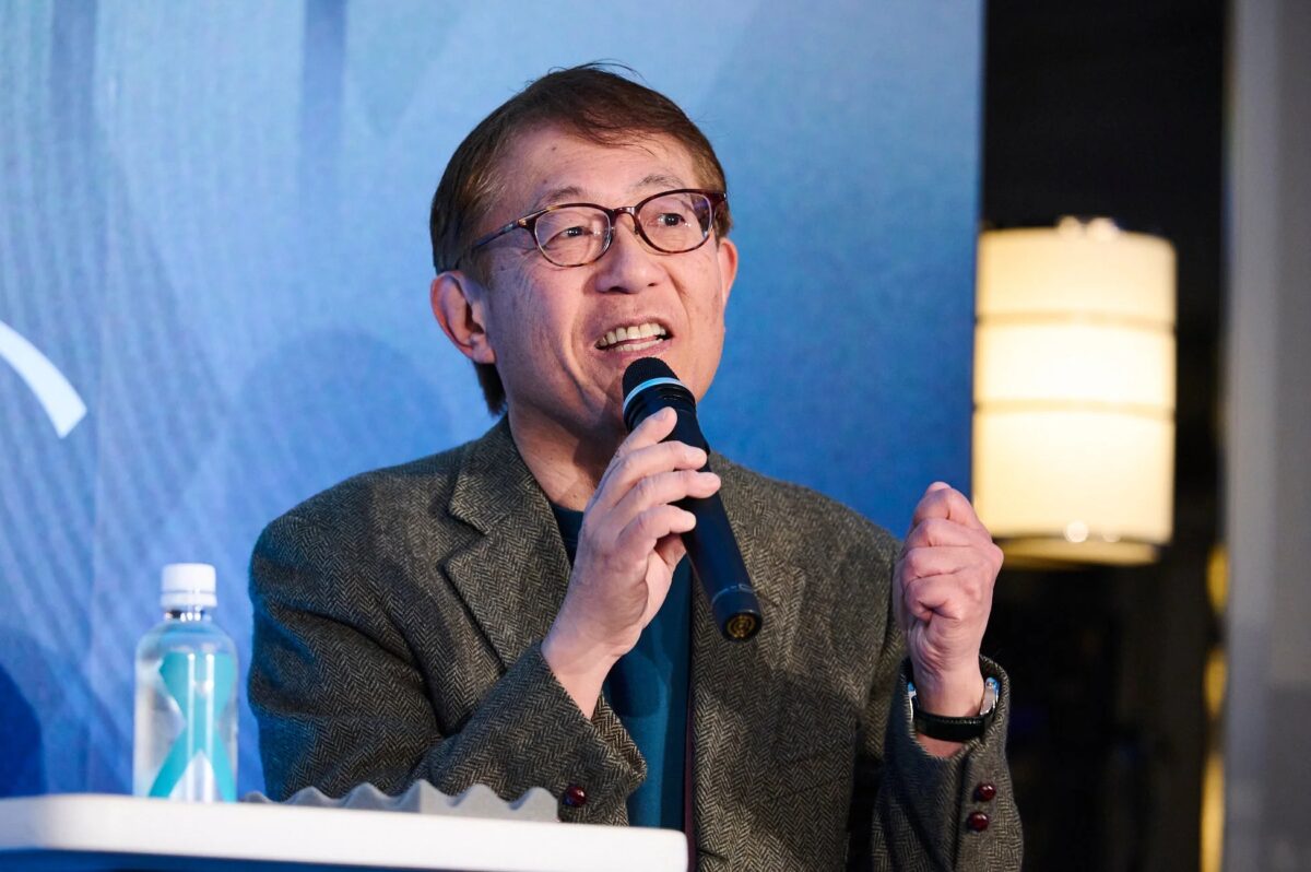 眼鏡をかけたアジア人男性がマイクを手に、会議やプレゼンテーションで熱心にスピーチしている様子。