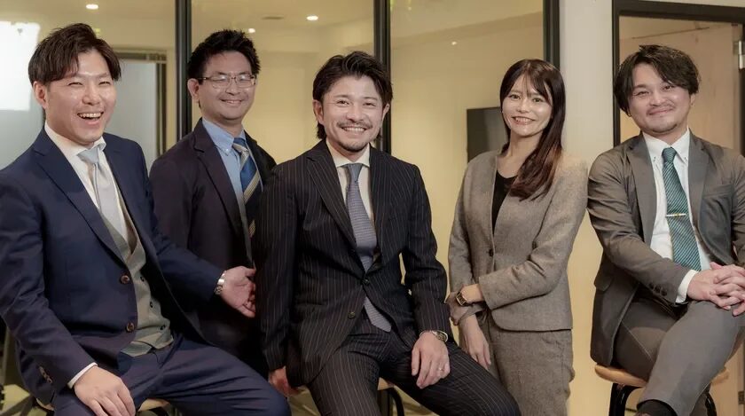 オフィスらしき空間で、スーツを着用した5人のビジネスパーソンが笑顔で座っています。