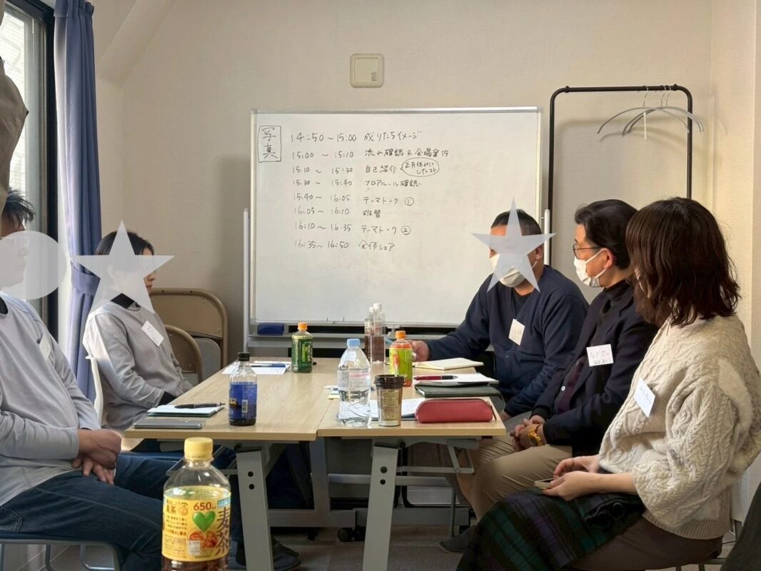 会議室で数人がテーブルを囲んで座り、ホワイトボードには「自己紹介」や「テーマトーク」といった議題が書かれたアジェンダが表示されています。飲み物が置かれ、活発な話し合いが行われている様子のミーティング風景です。