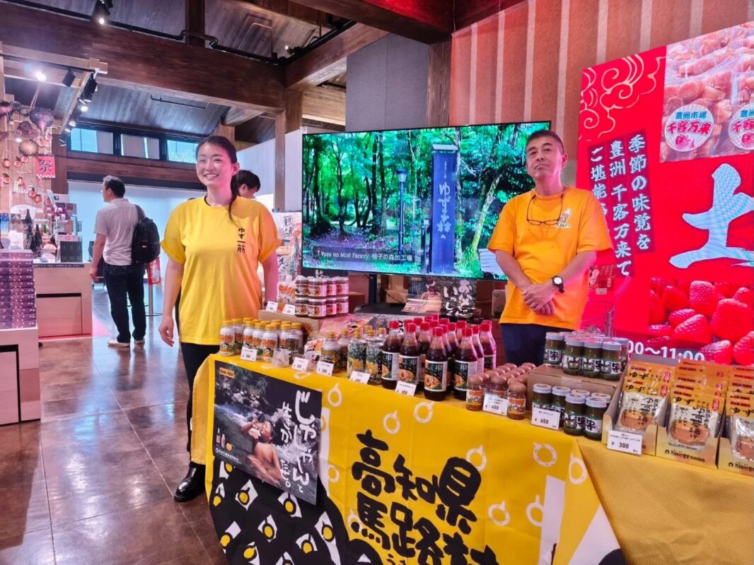 高知県馬路村のゆず製品を販売するイベントブースで、黄色のTシャツを着た男女スタッフが笑顔で立っています。