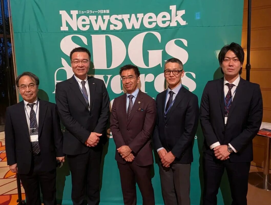 ニューズウィークSDGsアワードのイベントで、スーツ姿の男性5人が緑色のバナーを背景に並んで立っている集合写真です。