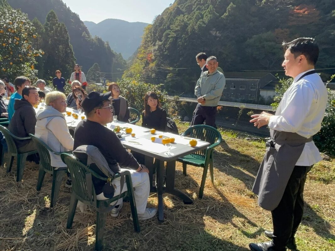 山々に囲まれた自然豊かな場所で、人々がテーブルを囲んで食事をしている様子です。