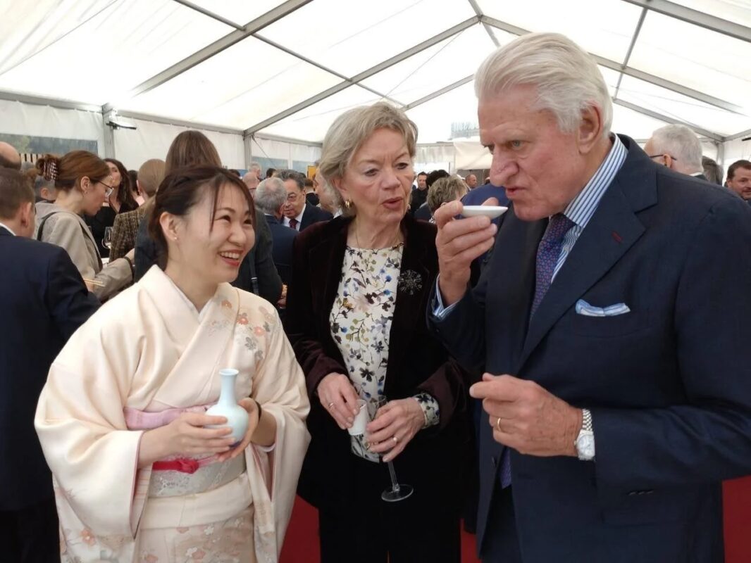 着物姿の日本人女性が笑顔で、小さな器から何かを試飲している外国人男性と交流している様子。