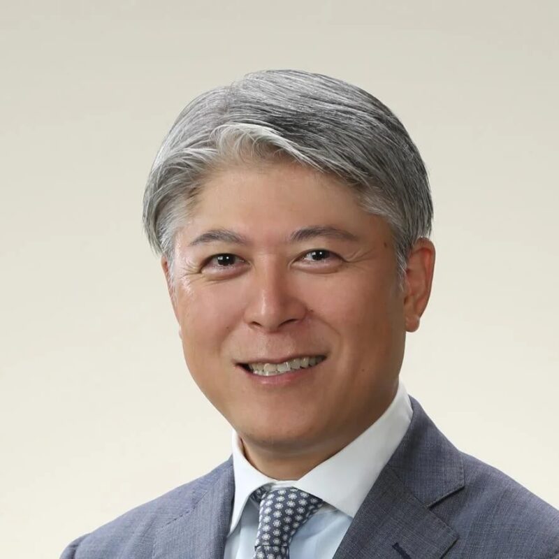 笑顔でカメラを見つめる中年のアジア人男性のポートレート。グレーの短髪で、水色のスーツと模様入りのネクタイを着用。親しみやすくプロフェッショナルな印象です。
