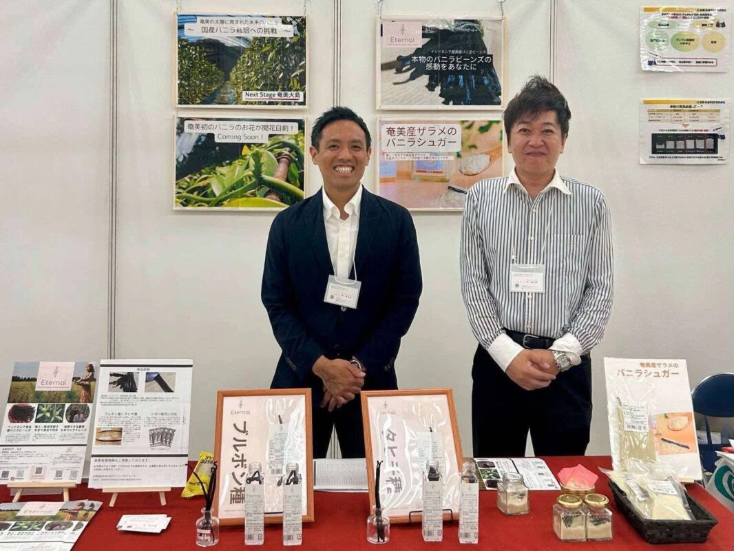 展示会ブースで2人の男性が、国産バニラ栽培やインドネシア産バニラビーンズ、奄美産ザラメのバニラシュガーといった「Eternal」ブランドのバニラ関連商品を展示・紹介しています。